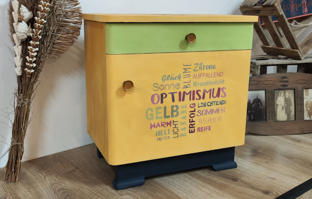 Ein Gelb und Grün angestrichenes Nachtschränkchen mit positven Wörtern zu Gelb auf der Tür